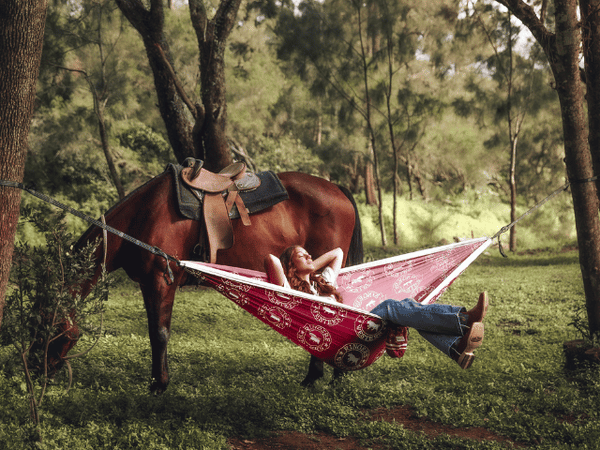 Red recycled hammock with straps suspended between trees, person relaxing underneath in forest setting.