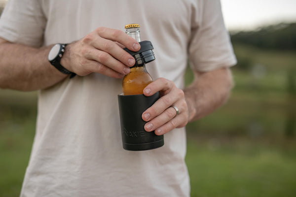 Black neoprene bottle cooler holding amber beer bottle with gold cap, worn on outdoor setting