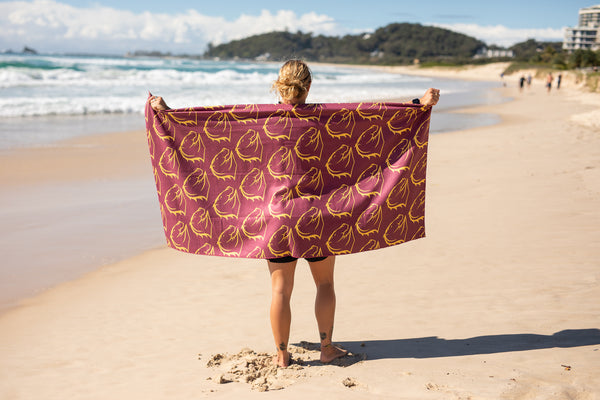 Burgundy and gold leaf-patterned recycled beach towel held open by person on sandy beach