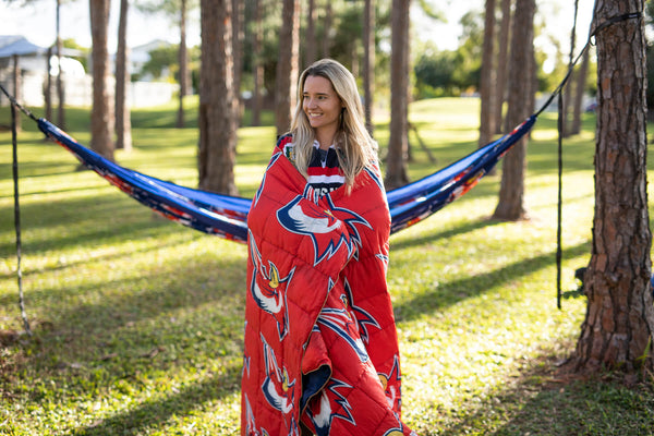 Red NRL Roosters sustainable down puffy blanket with navy and white bird logo design, draped around woman in hammock