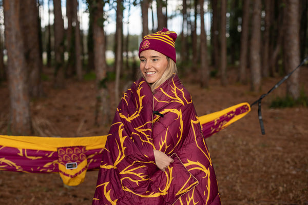 Burgundy and gold NRL puffy blanket with geometric pattern, worn as a wrap with matching beanie in forest setting.