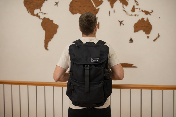 A man viewed from behind wearing a black Nake backpack, standing in front of a wall with a wooden world map.
