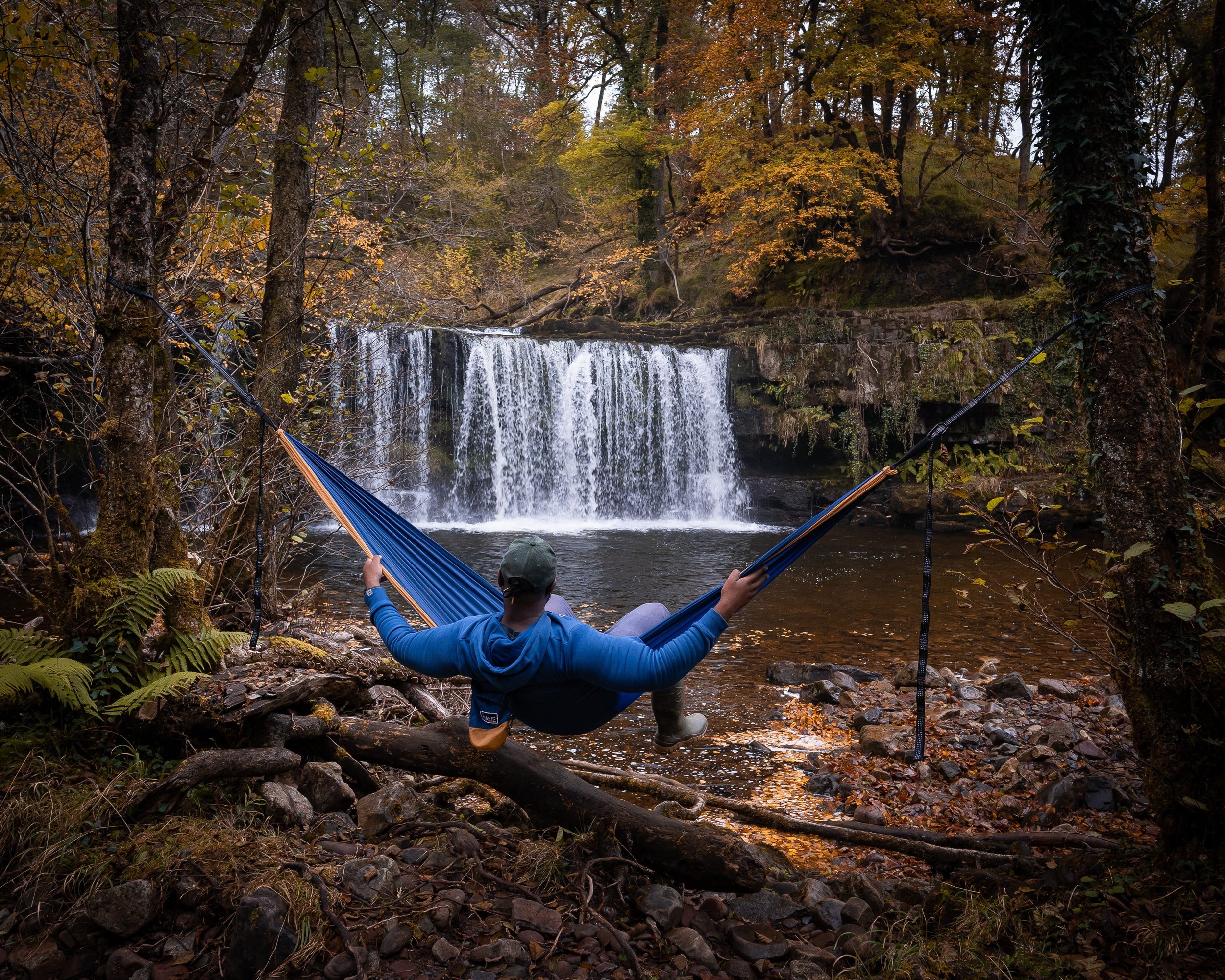 Person relaxing in a Nakie River Blue hammock fronting a waterfall in a forest.