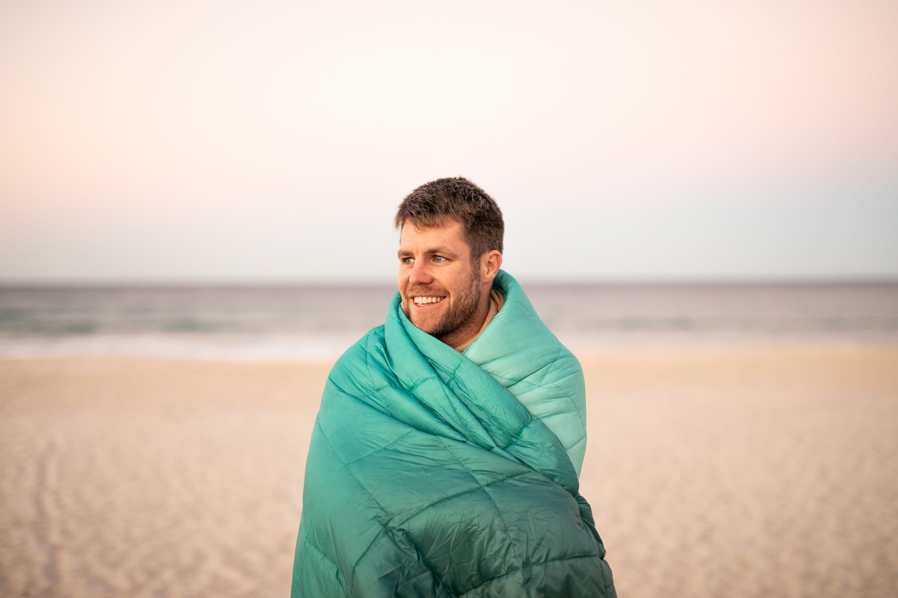 Man standing on the beach wrapped in a Nakie Horizon Green puffy blanket
