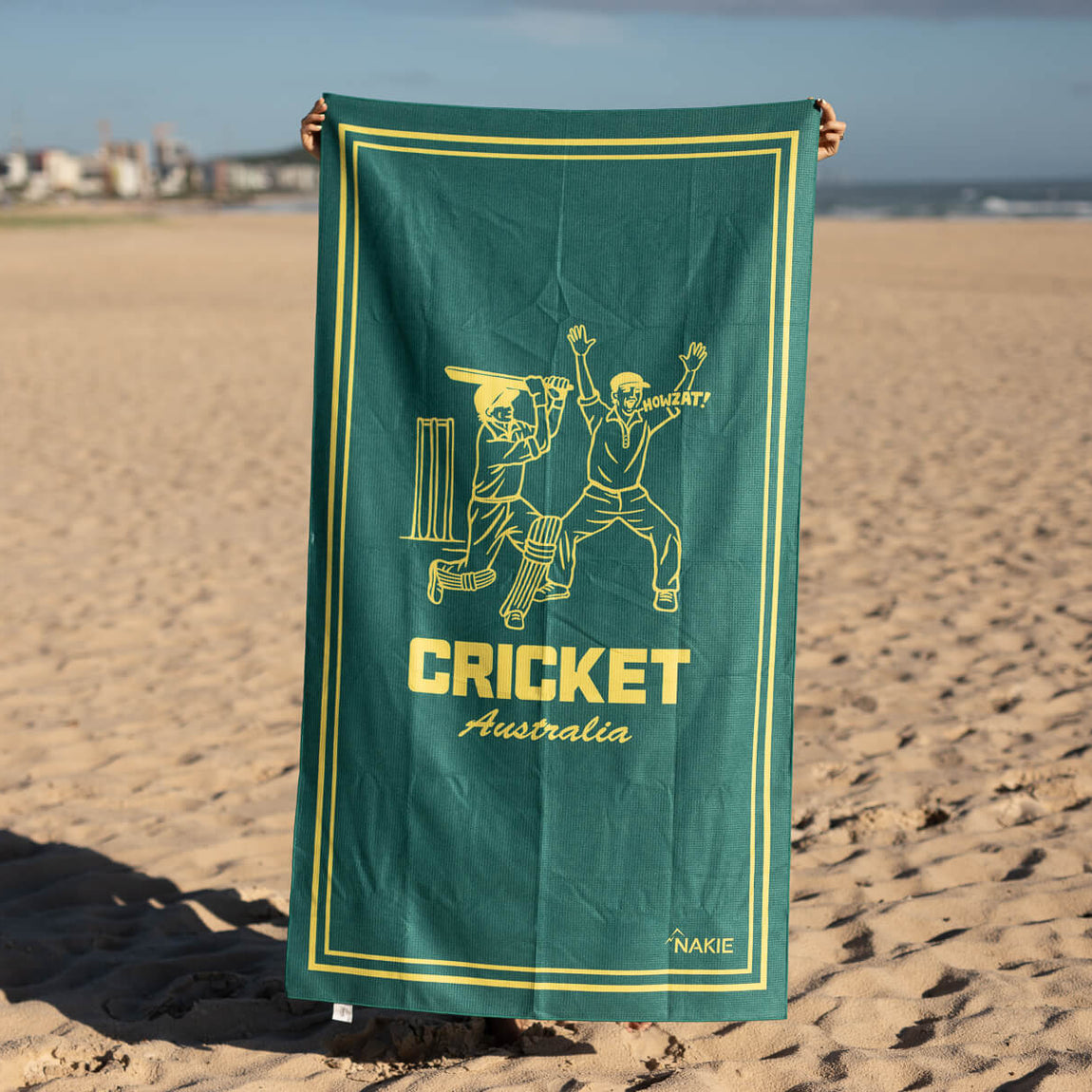 Green and gold Cricket Australia beach towel with illustrated cricketers and wickets, displayed on sand