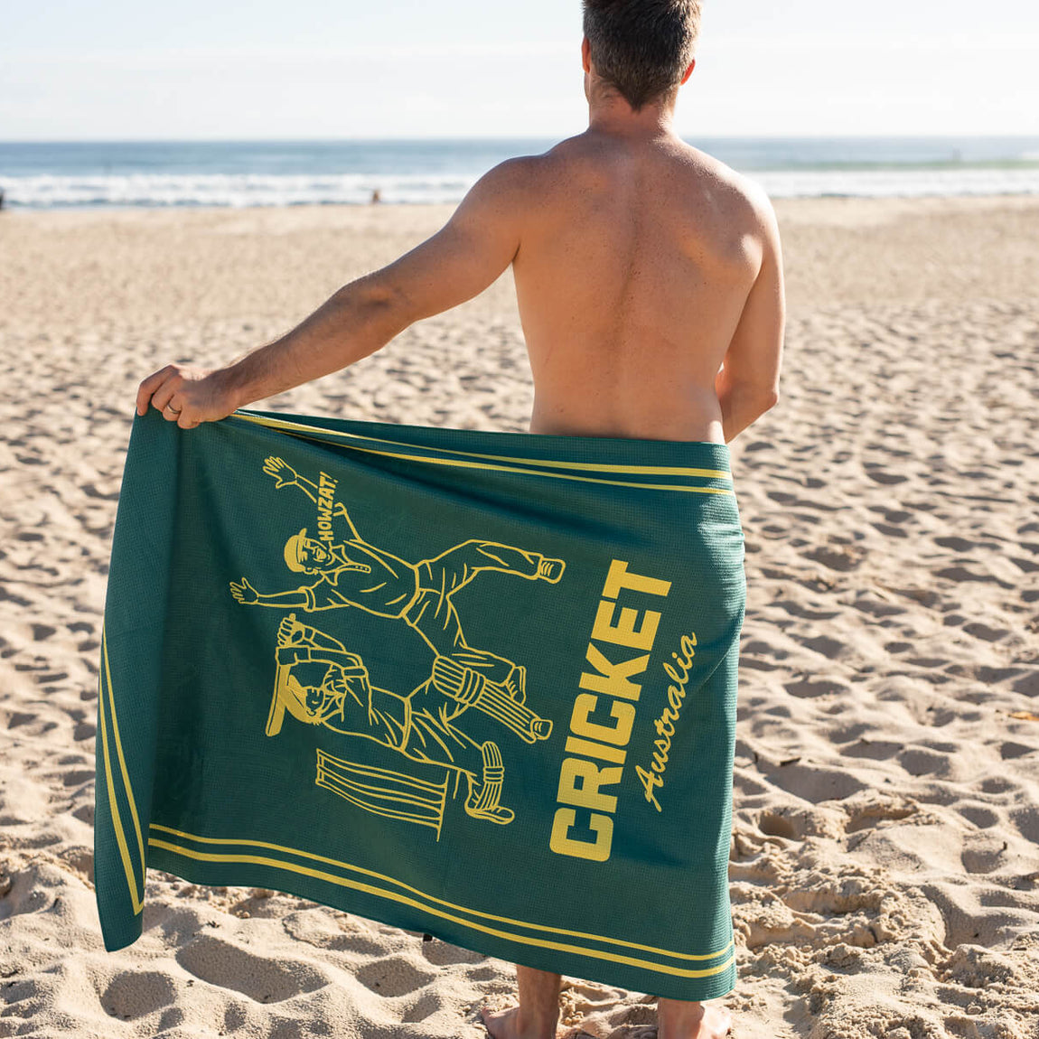 Teal beach towel with yellow cricket player graphic and CRICKET Australia text, displayed on sand at beach