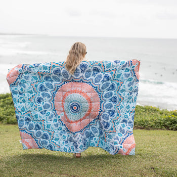 Blue and coral mandala-print sustainable down puffy blanket held up outdoors overlooking ocean.