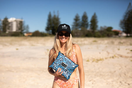 Blue and grey recycled sand-free beach towel with abstract wave pattern, held on beach by woman wearing cap and sunglasses