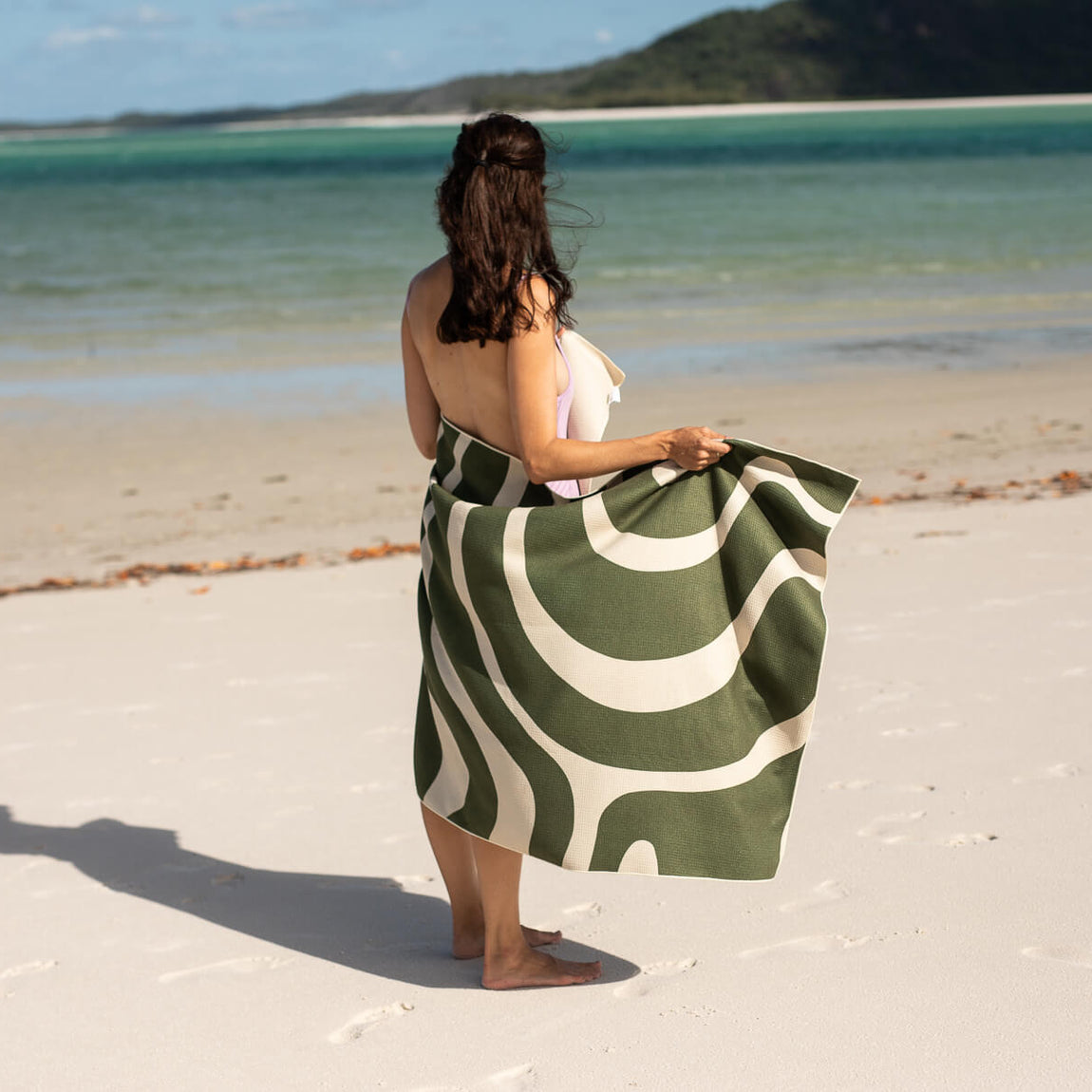 Eucalyptus green beach towel with cream wavy stripes, held by woman on sandy beach overlooking turquoise water