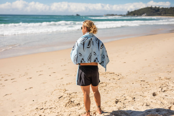 Light blue patterned beach towel with dark leaf motif, draped over woman's shoulders on sand.