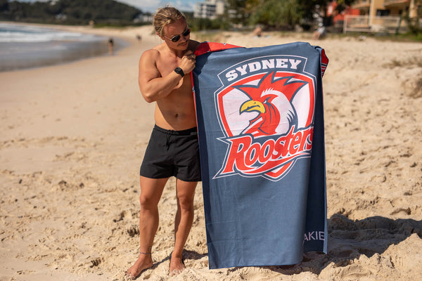 Sydney Roosters NRL beach towel in navy with red rooster logo, displayed on sand at beach