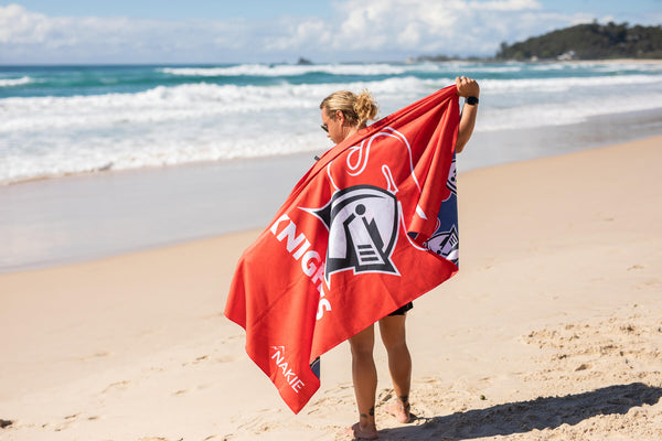 Red Knights NRL beach towel with white graphic logo and text, displayed on sandy beach with ocean waves