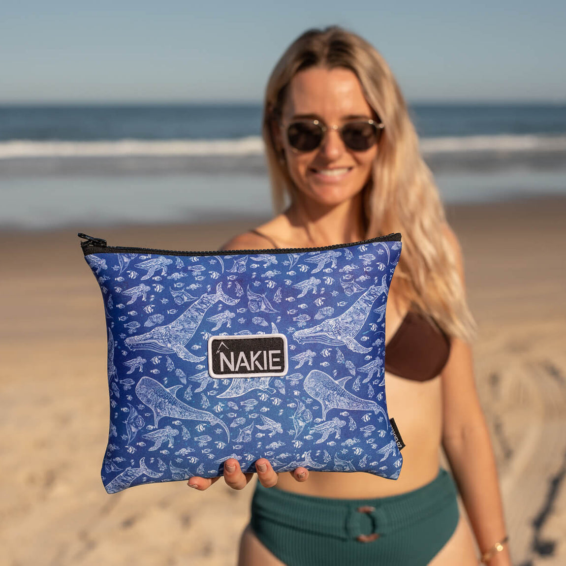 Blue recycled beach towel with white marine and fish pattern, held up on sandy beach