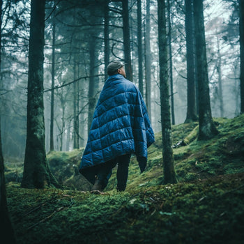 Image of a man wrapped in Nakie's Deep Ocean Blue Sustainable Down Puffy Blanket while standing in a campsite, enjoying warmth and sustainability during outdoor adventures.