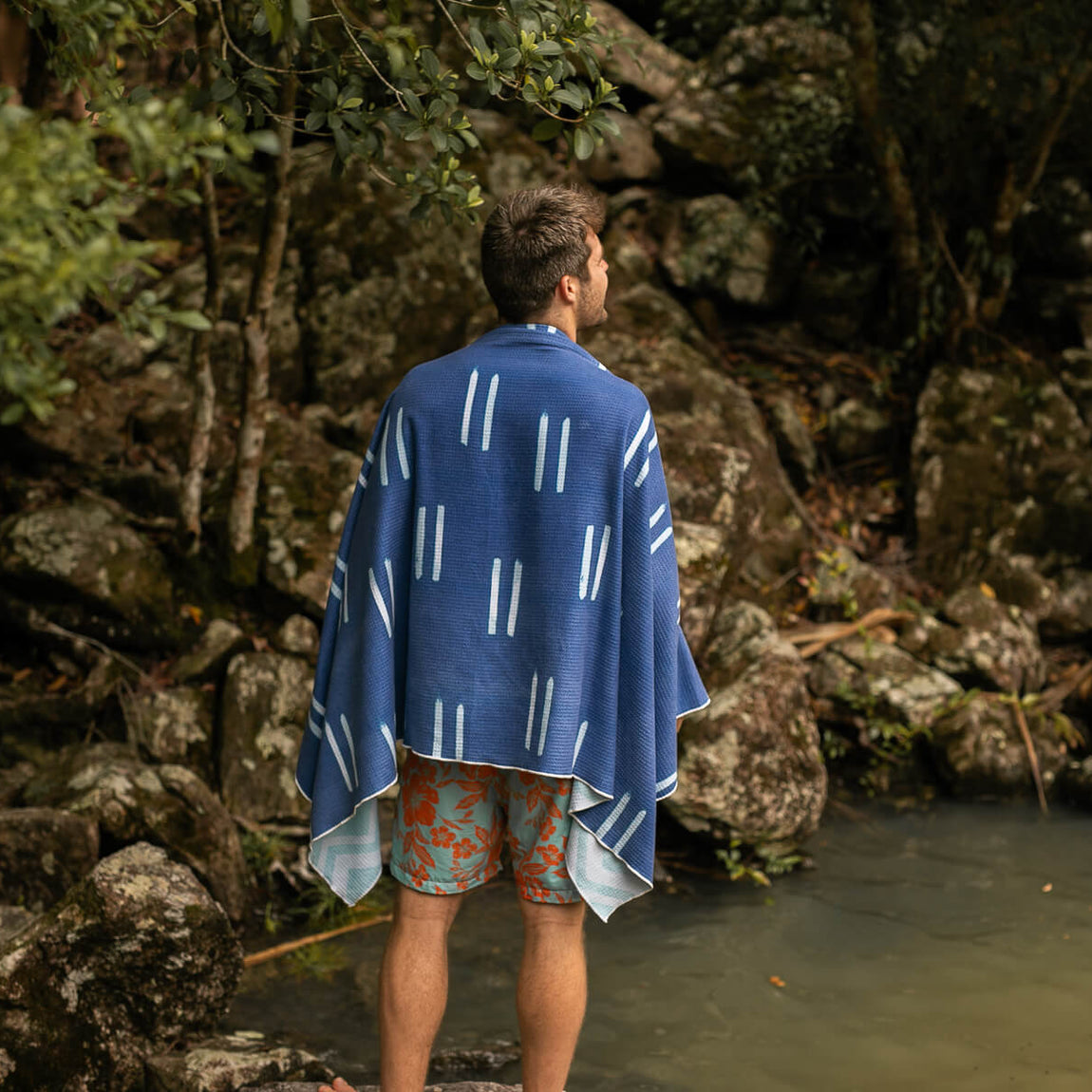 Blue recycled sand-free beach towel with white geometric stripe pattern, draped over shoulders by water's edge